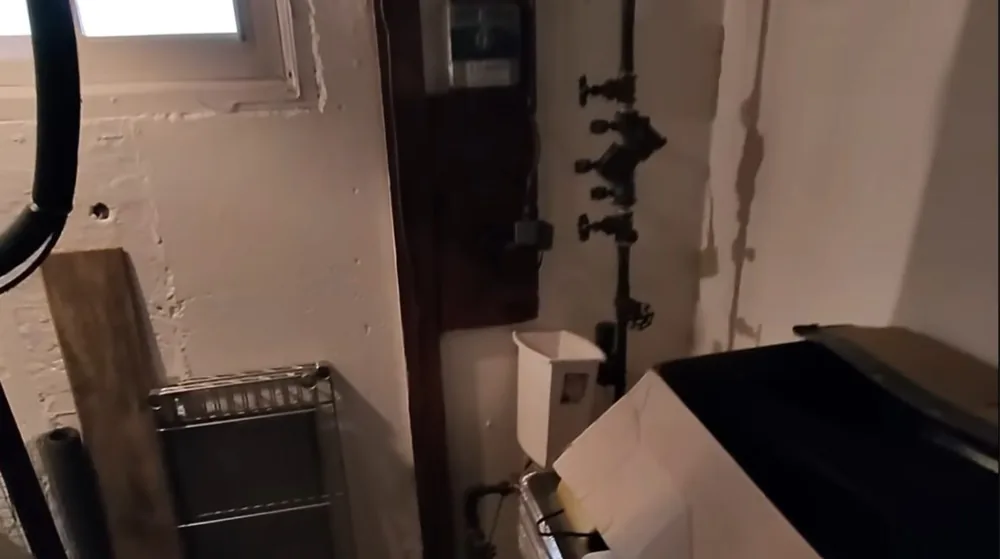 Basement wall with exposed pipes and shutoff valves above a dark appliance and a white box
