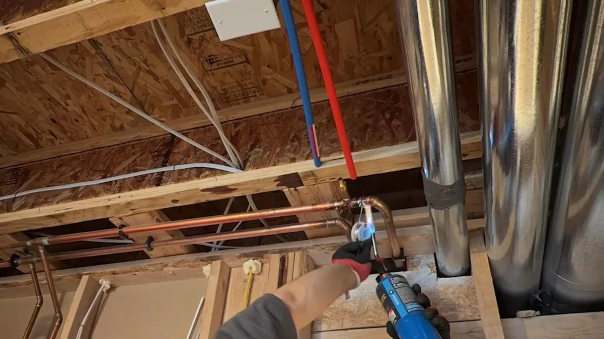 Ceiling joists with red and blue PEX lines and metal ductwork overhead, a hand holding a tool at one connection