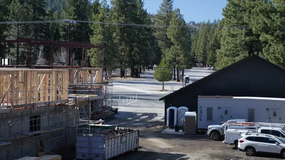 Construction site beside a road with an open trench, scaffolding and a dumpster, with pine trees behind