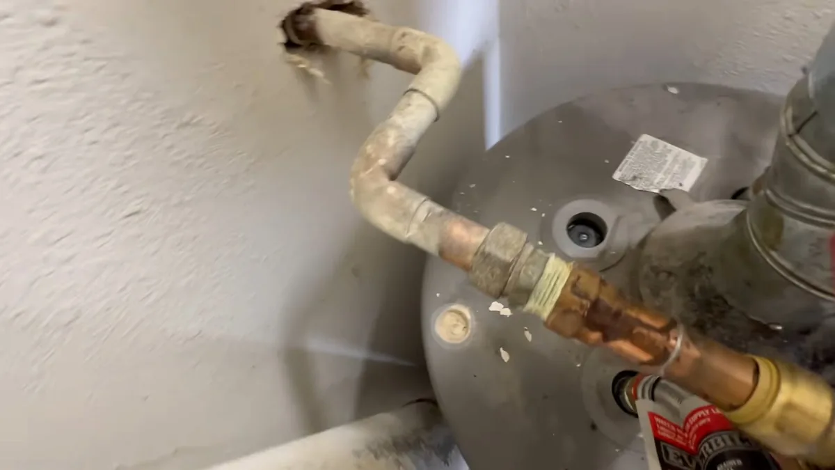 Copper and brass fittings on top of a gray tank water heater, with a wall behind it