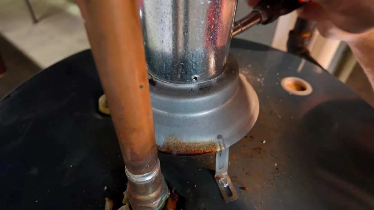 Copper pipe and a galvanized vent on top of a black water heater, with a torch flame and a hand at the top edge