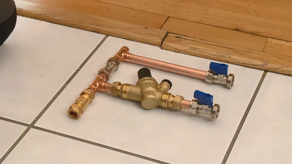 Copper pipe sections, a brass pressure regulator and two blue-handled shutoff valves laid out on white floor tile