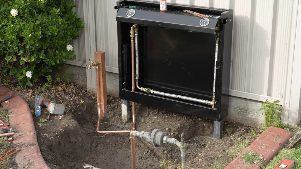 Copper service pipes running from a water meter and shutoff valves to a black outdoor equipment cabinet beside a white fence