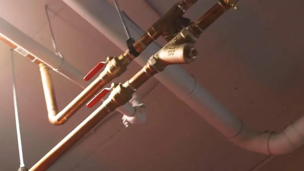 Copper water supply pipes with brass ball valves running along an exposed ceiling beside white drain pipe