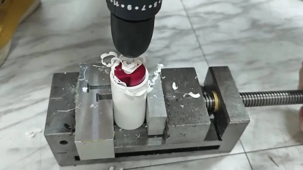 Drill bit cutting into a white PVC fitting held in a metal vise on a marble surface