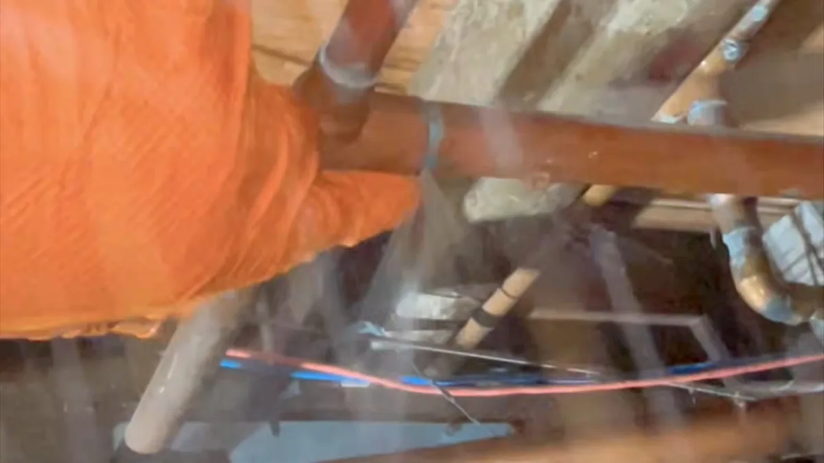 Gloved hand holding a copper pipe inside a framed wall as water sprays past