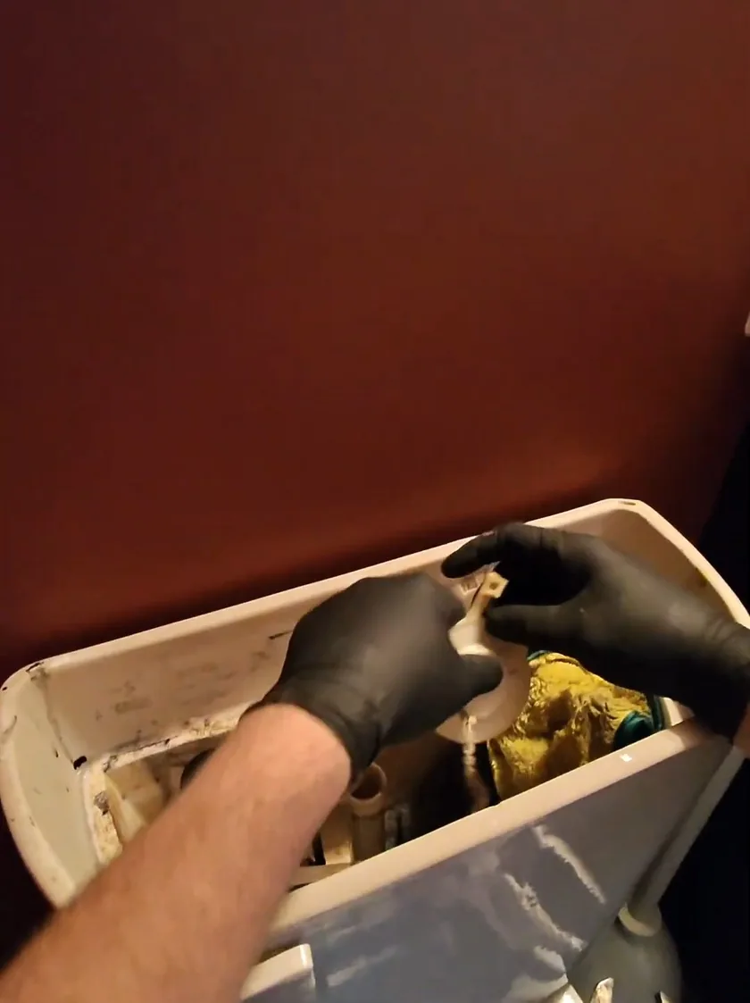 Gloved hand reaching into an open toilet tank to hold a repair part among the fill valve parts