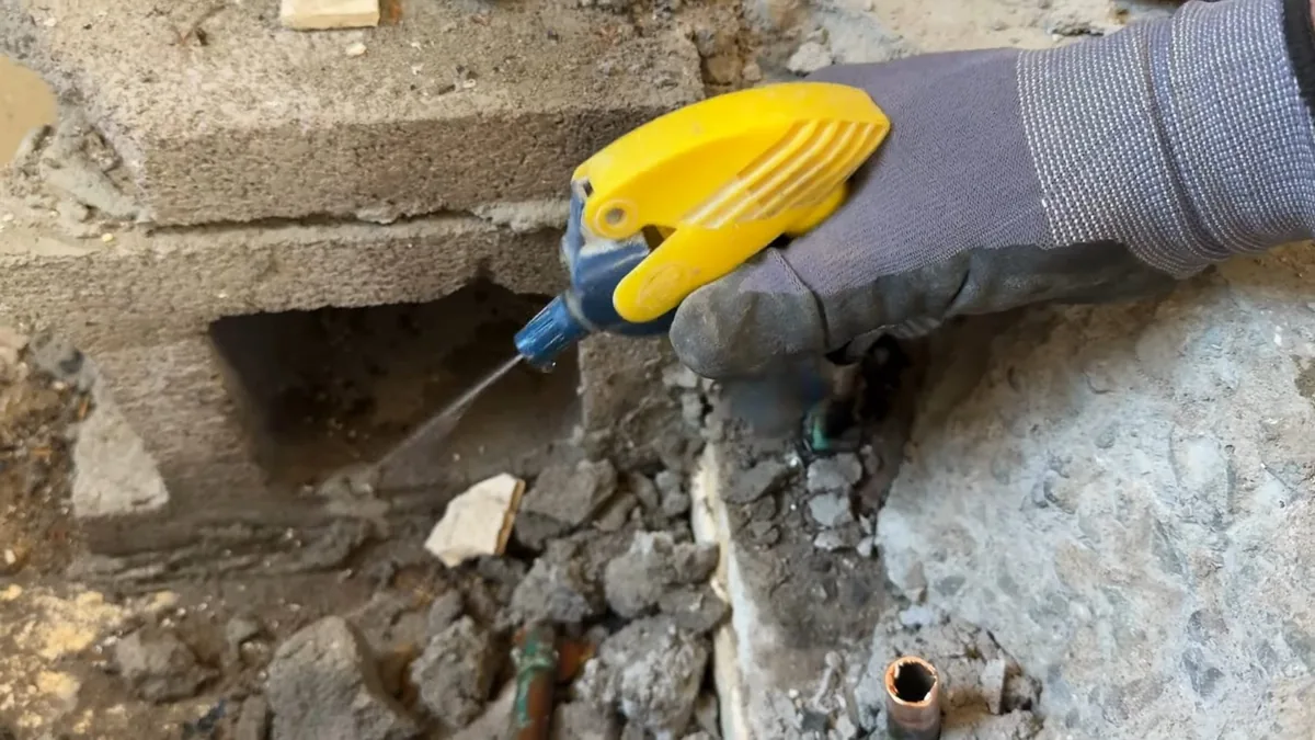 Gloved hand using a yellow tool at a broken section of concrete to reach a pipe underneath