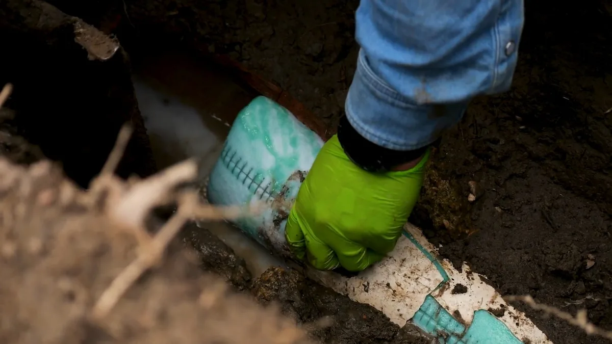 Gloved hand working a liner into an underground pipe in a dirt trench