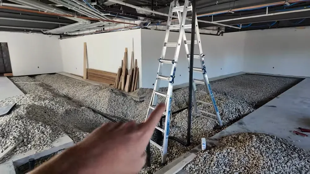 Gravel bed laid in trench lines across the floor of an unfinished commercial space with a ladder and lumber behind