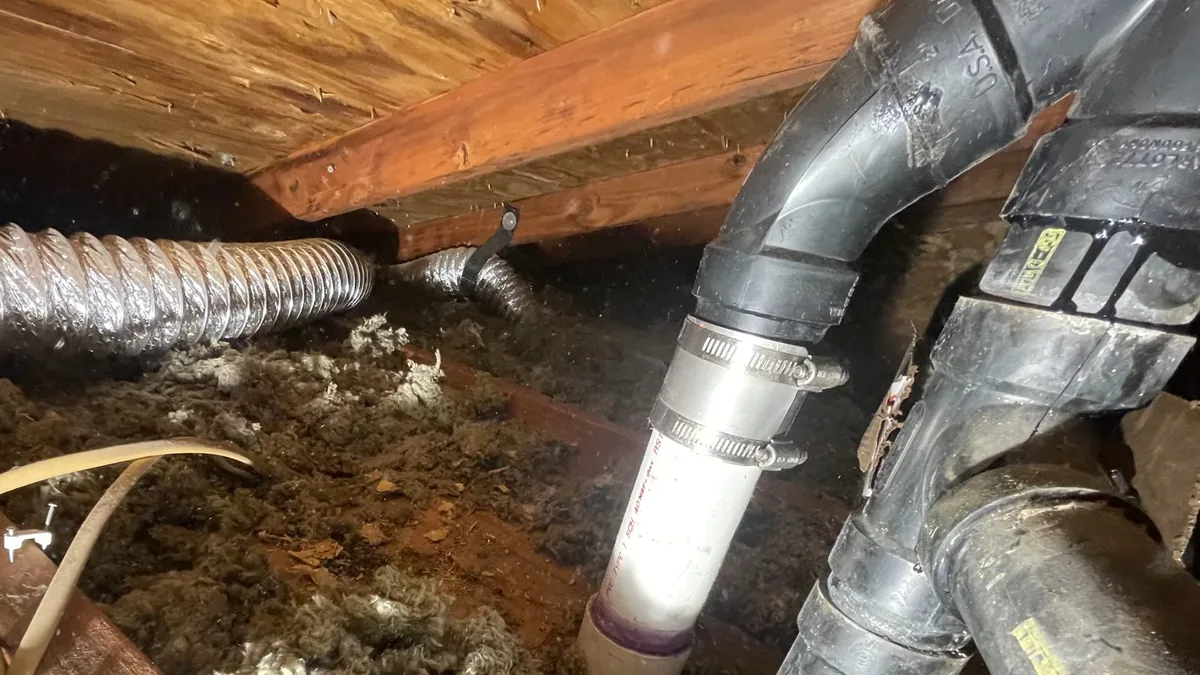 Gray drain elbow and a length of flexible duct under wooden joists in a crawl space