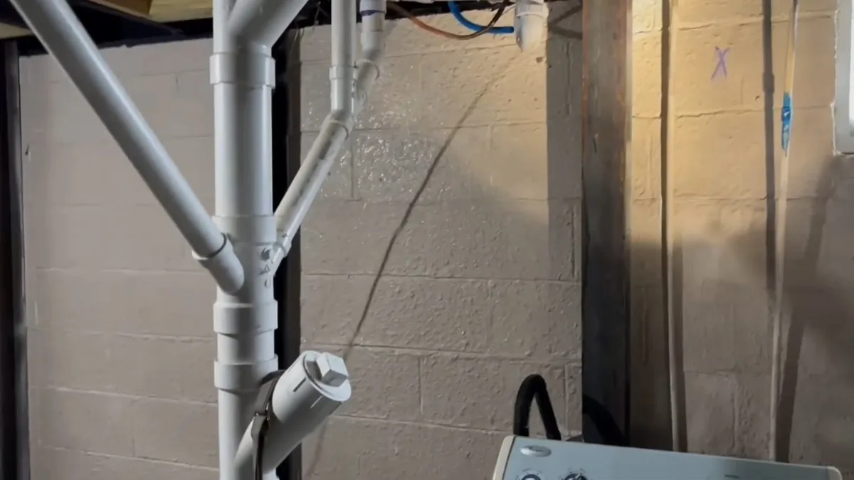 Gray PVC drain pipes branching against a concrete block wall, with a cleanout fitting at the lower pipe
