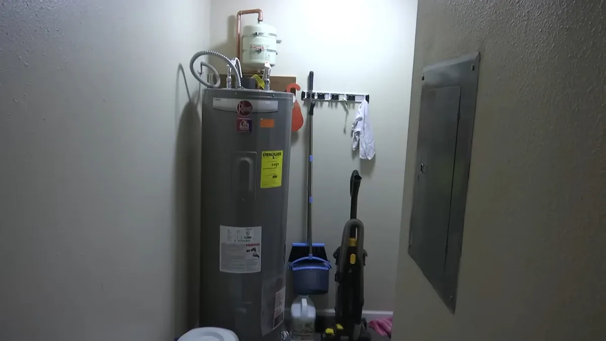 Gray tank water heater standing in a closet with a mop and a vacuum beside it and a panel on the wall