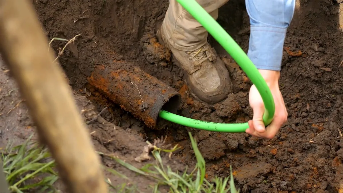 Green flexible tube fed into the open end of a rusted pipe in a dug-out section of yard