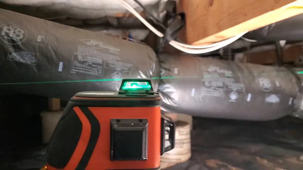 Green laser level sitting on the floor beside insulated duct in a crawl space