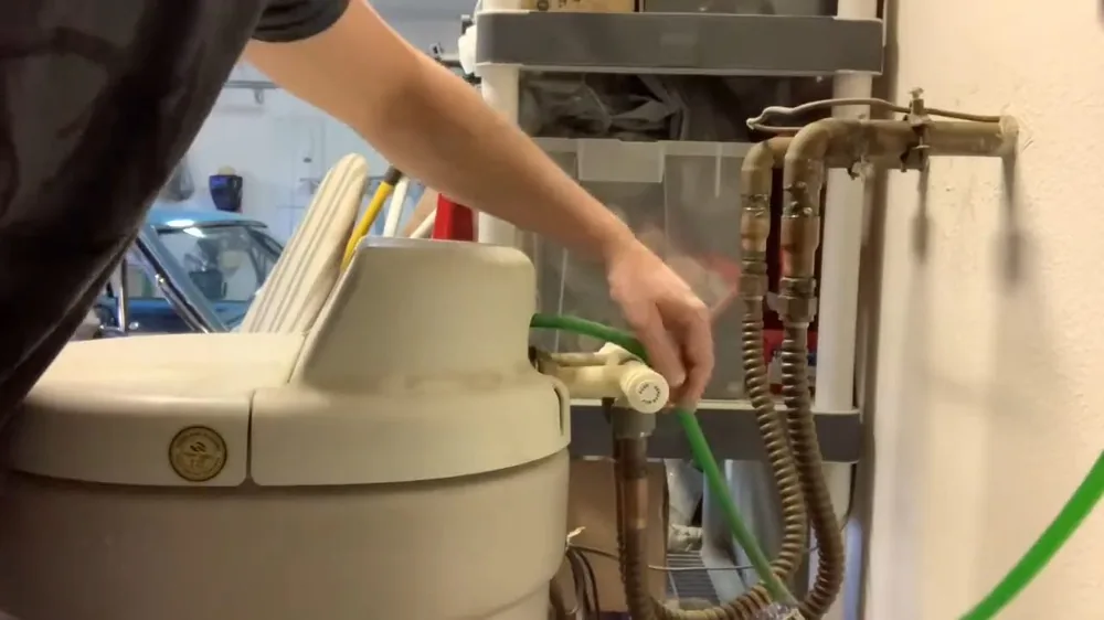 Hand adjusting a green hose connection on a beige water softener tank beside copper plumbing