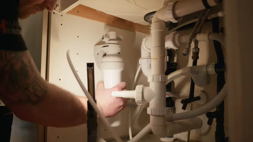 Hand holding a white drain trap section under a sink among tangled white drain and supply pipes