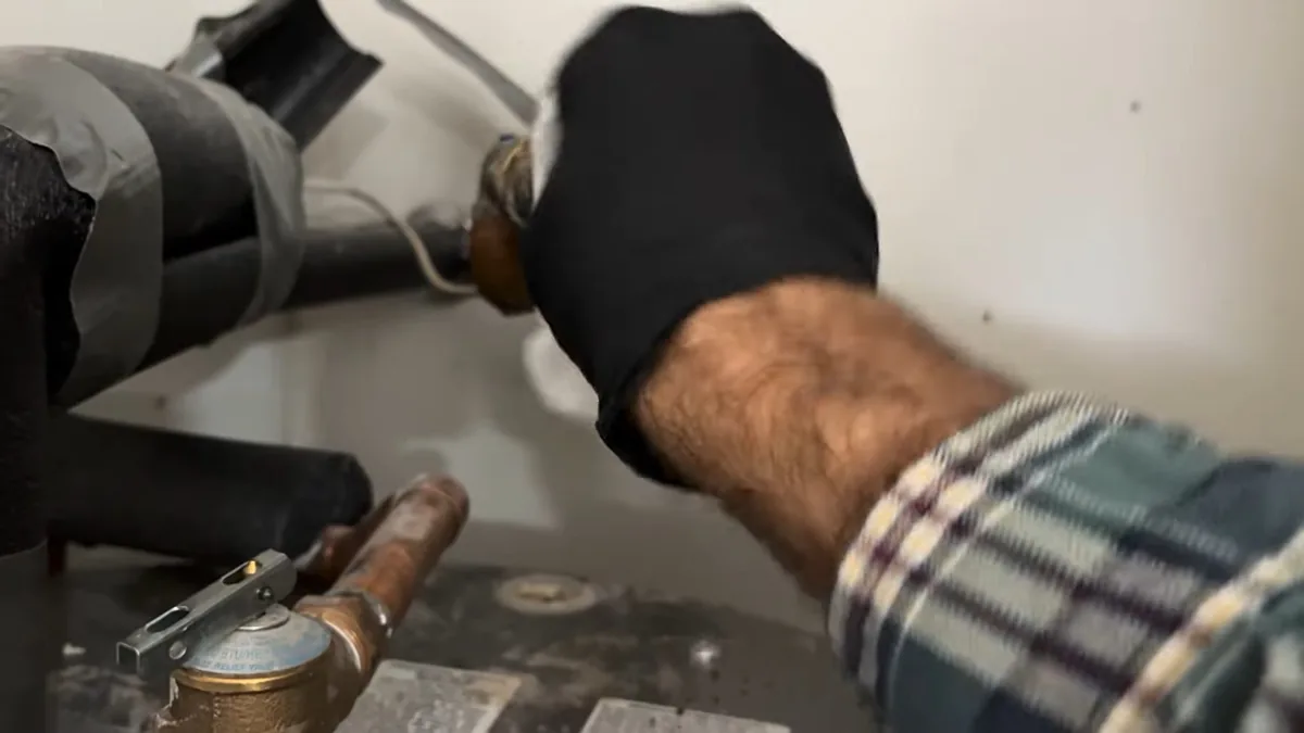 Hand in a black glove and plaid sleeve working a brass fitting near insulated pipe