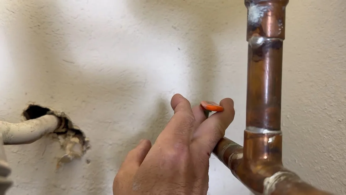 Hand marking a copper pipe repair beside a hole cut in a painted wall
