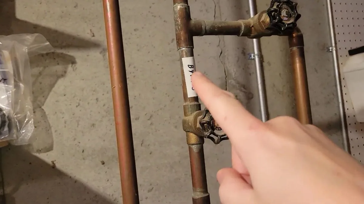 Hand pointing at a brass shutoff valve on copper pipes mounted on a concrete wall