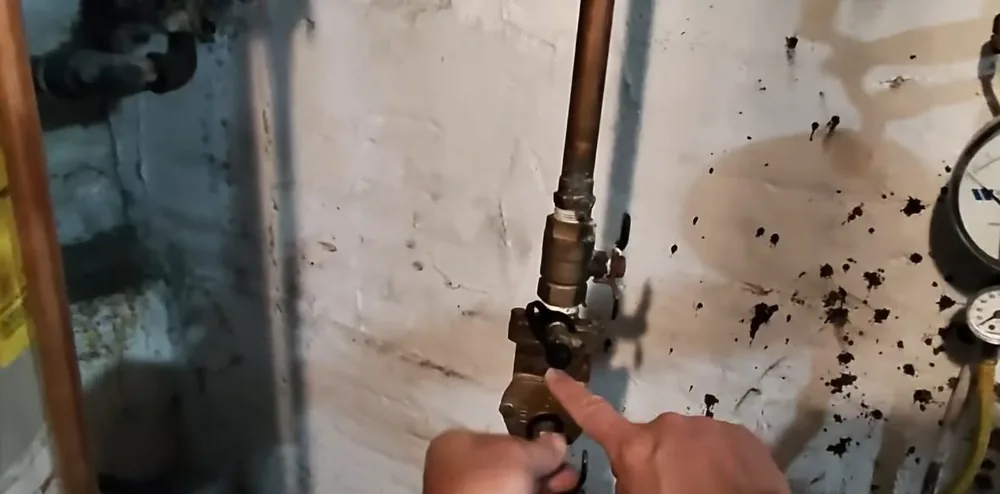 Hand pointing at a brass valve on a copper pipe beside pressure gauges on a stained basement wall