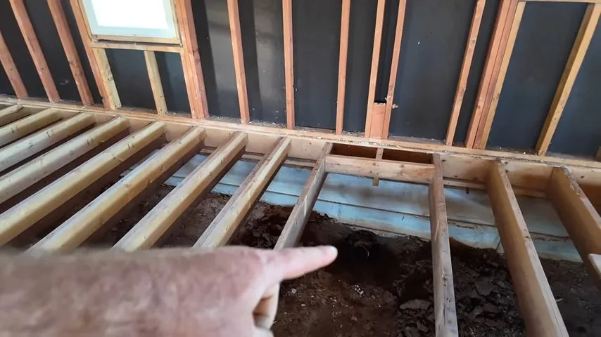 Hand pointing into an exposed floor frame where joists are open above bare soil