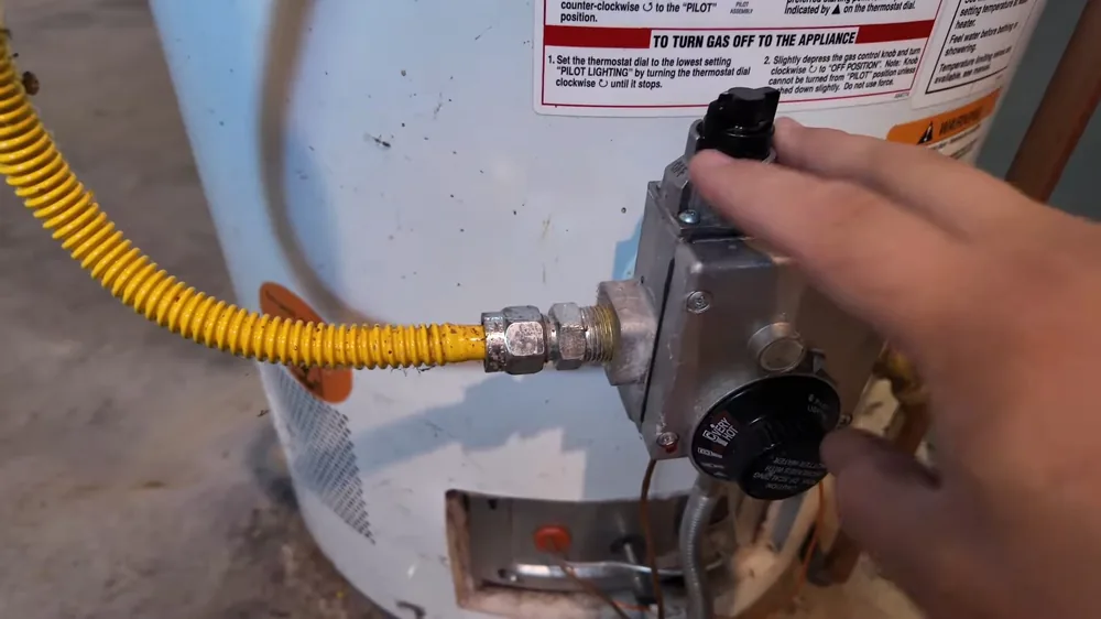 Hand turning the gas control on a water heater, with a yellow gas line and warning label beside it