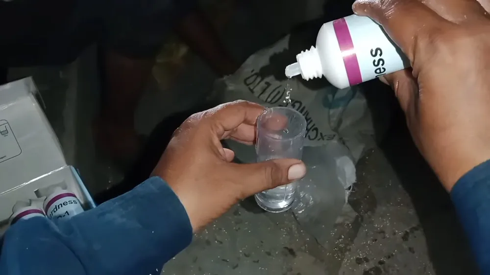 Hands adding drops from a small bottle to a glass sample of tap water on a dark surface
