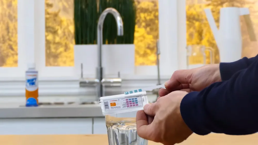 Hands dipping a water test strip into a glass of tap water beside a kitchen window