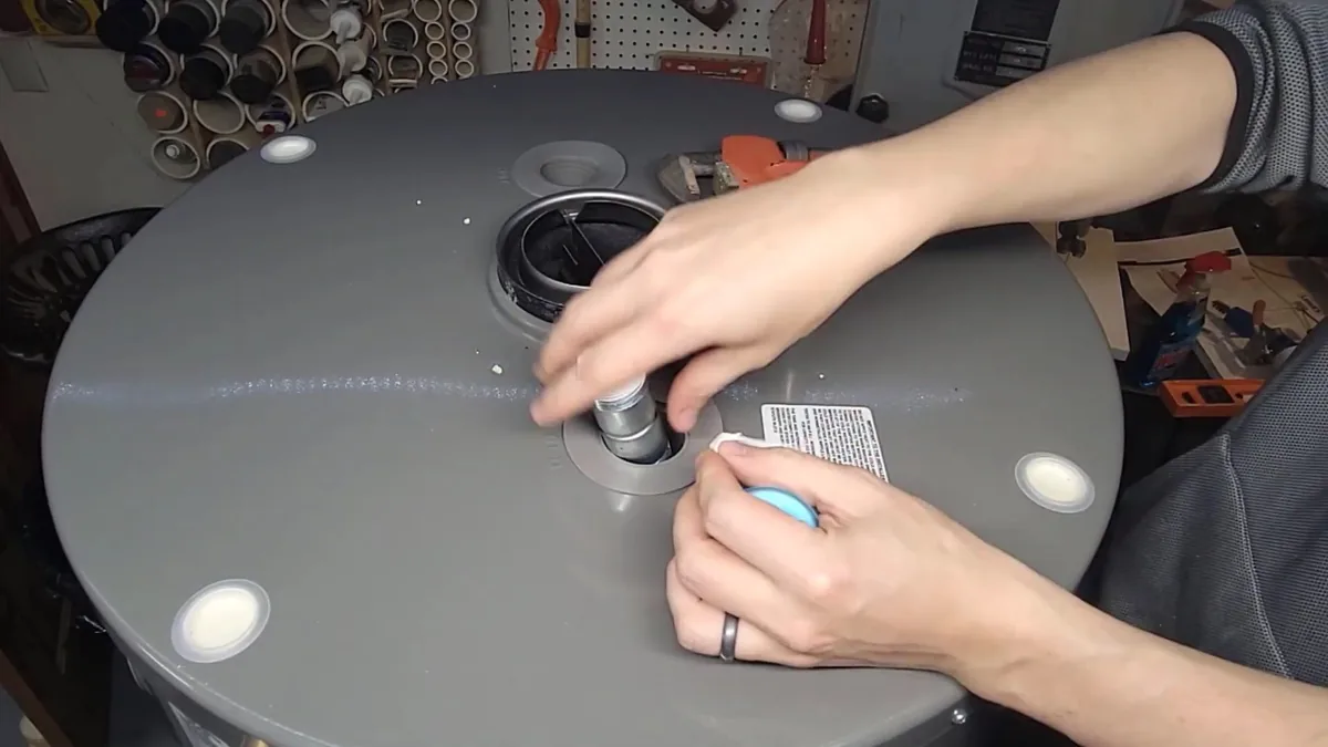 Hands fitting a part into a port on top of a gray tank water heater, with a pipe wrench behind