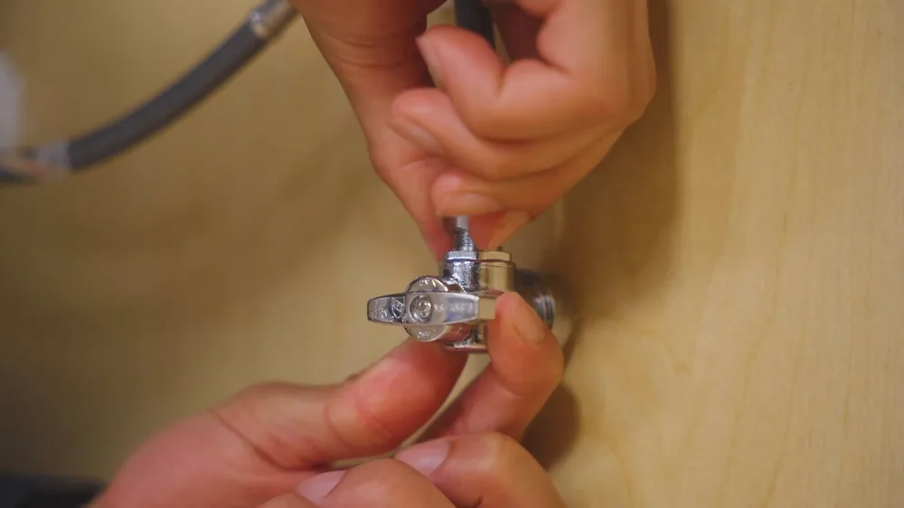 Hands tightening a chrome angle stop valve onto a supply line against a wood panel