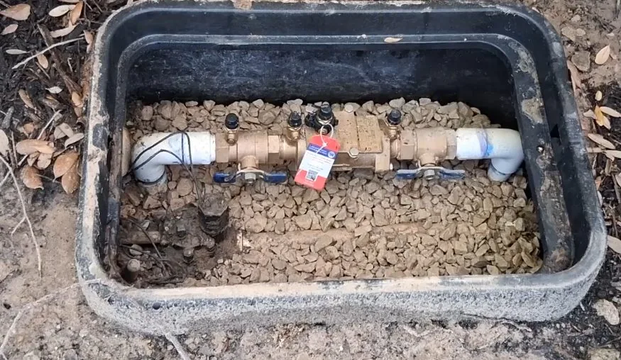 Open in-ground box holding a brass pipe assembly with two shutoff valves and a red tag, set in gravel between white pipe ends