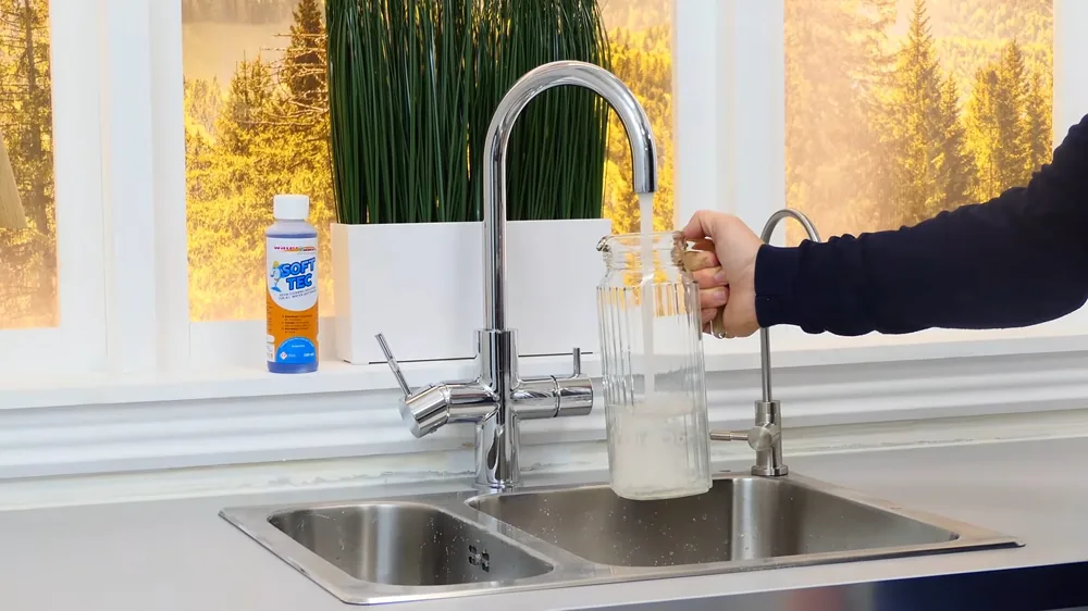 Kitchen faucet with a filter spout filling a glass pitcher at a double sink, with a bottle by the window