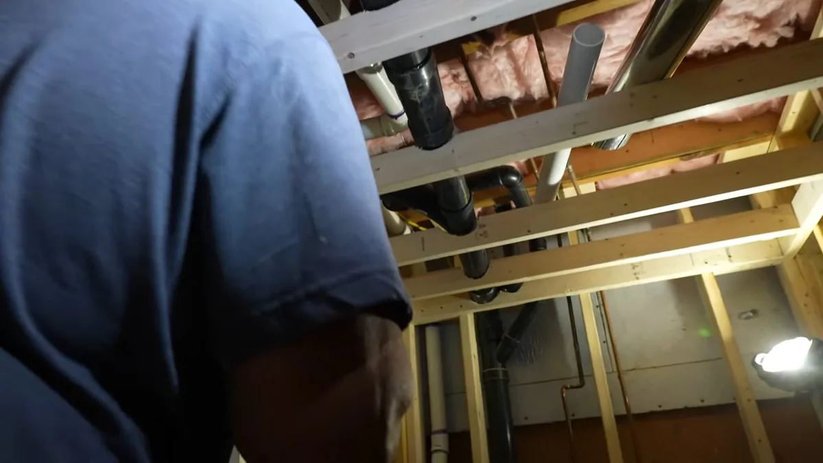 Man crouching at open framing in a basement, with white PVC drain lines running overhead
