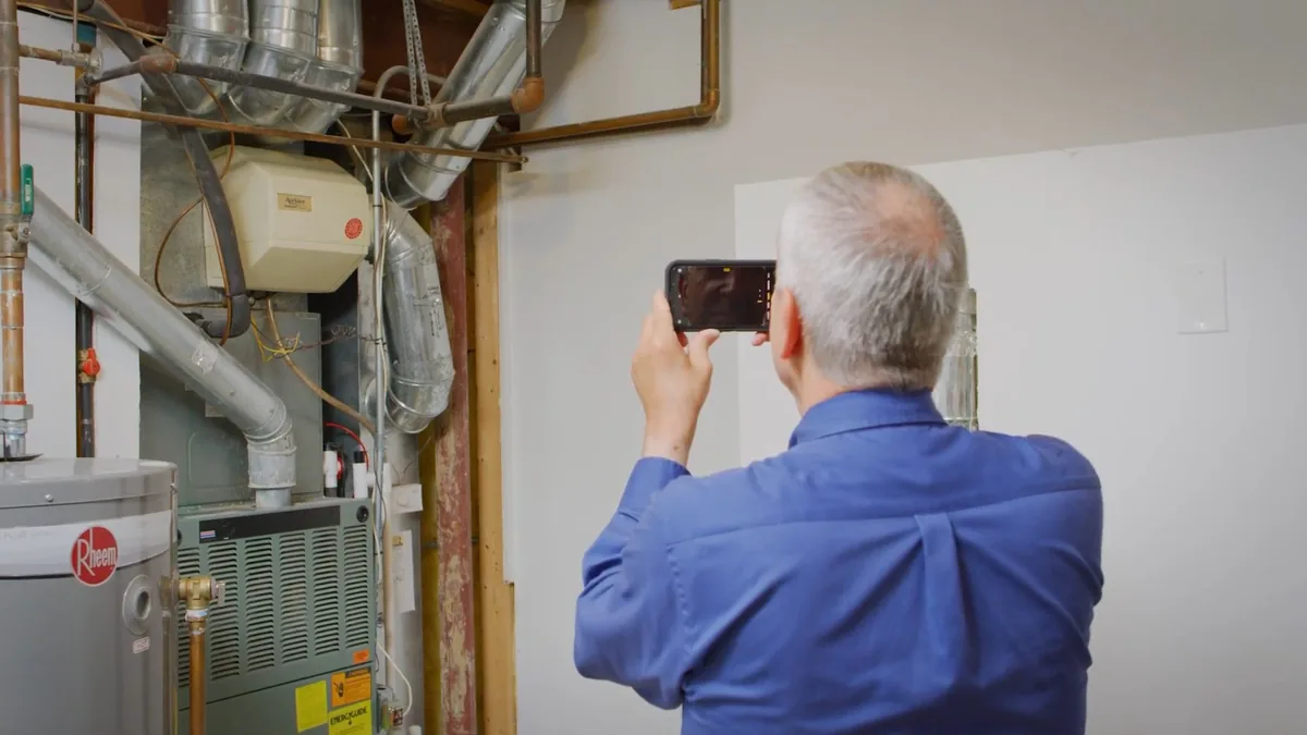 Man in a blue shirt photographing a water heater and furnace closet with his phone