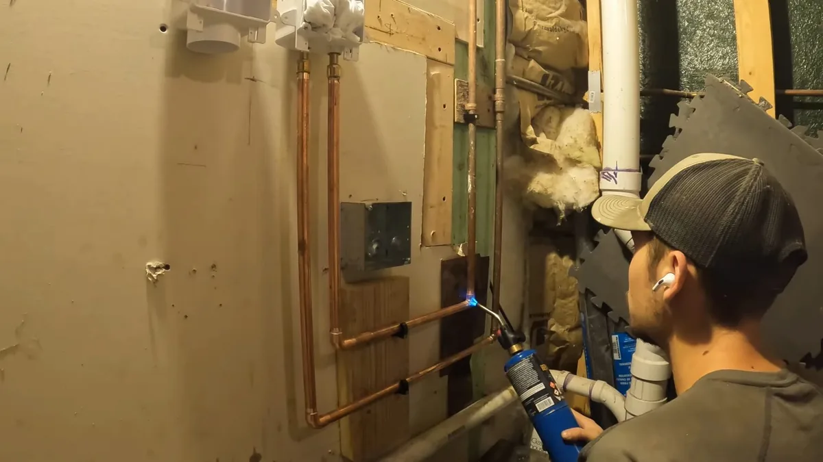 Man in a cap soldering copper lines on a basement wall with a torch, with a white drain pipe beside him