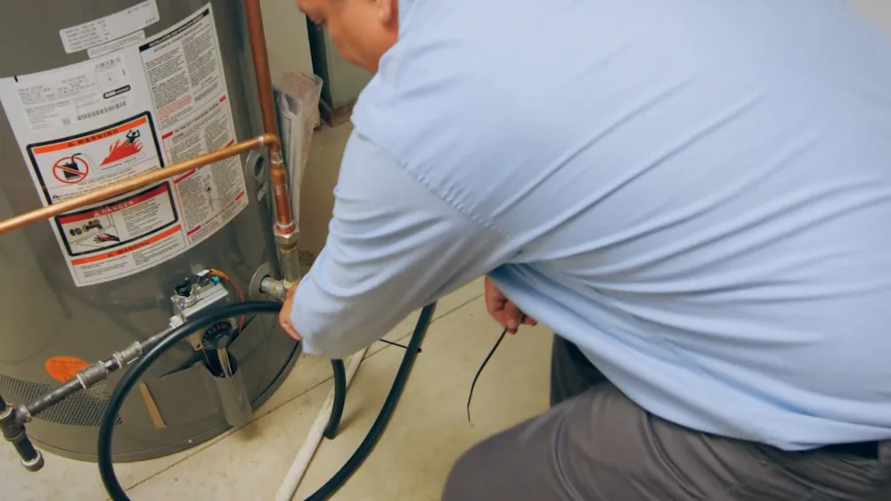 Man in a light blue shirt kneeling at a tank water heater with a black hose and warning labels visible