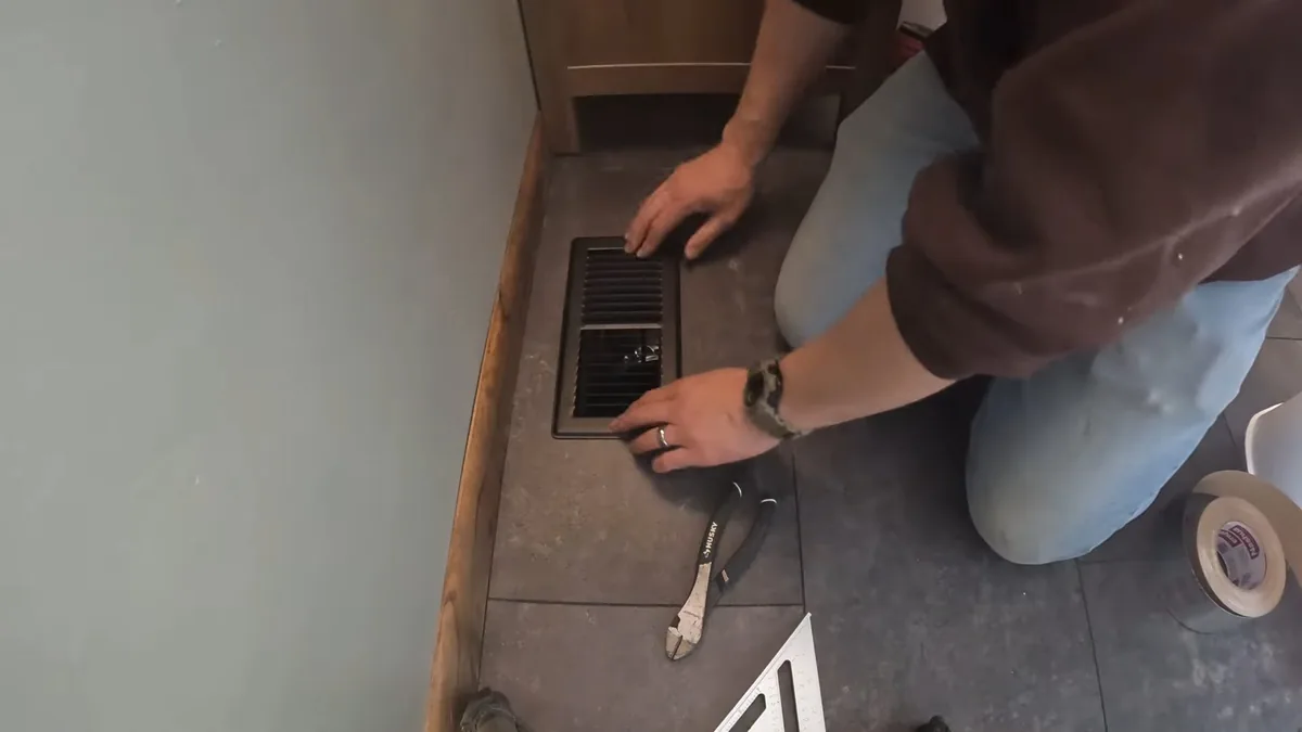 Man kneeling on a tile floor reaching toward a floor vent with a wrench lying beside him