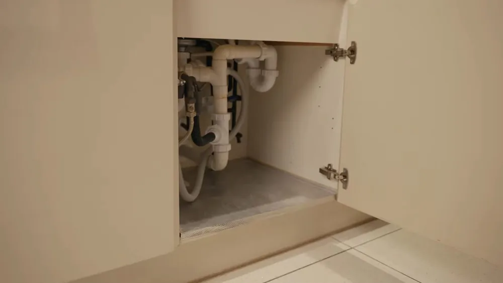 Open kitchen cabinet under a sink showing white drain pipes and supply lines