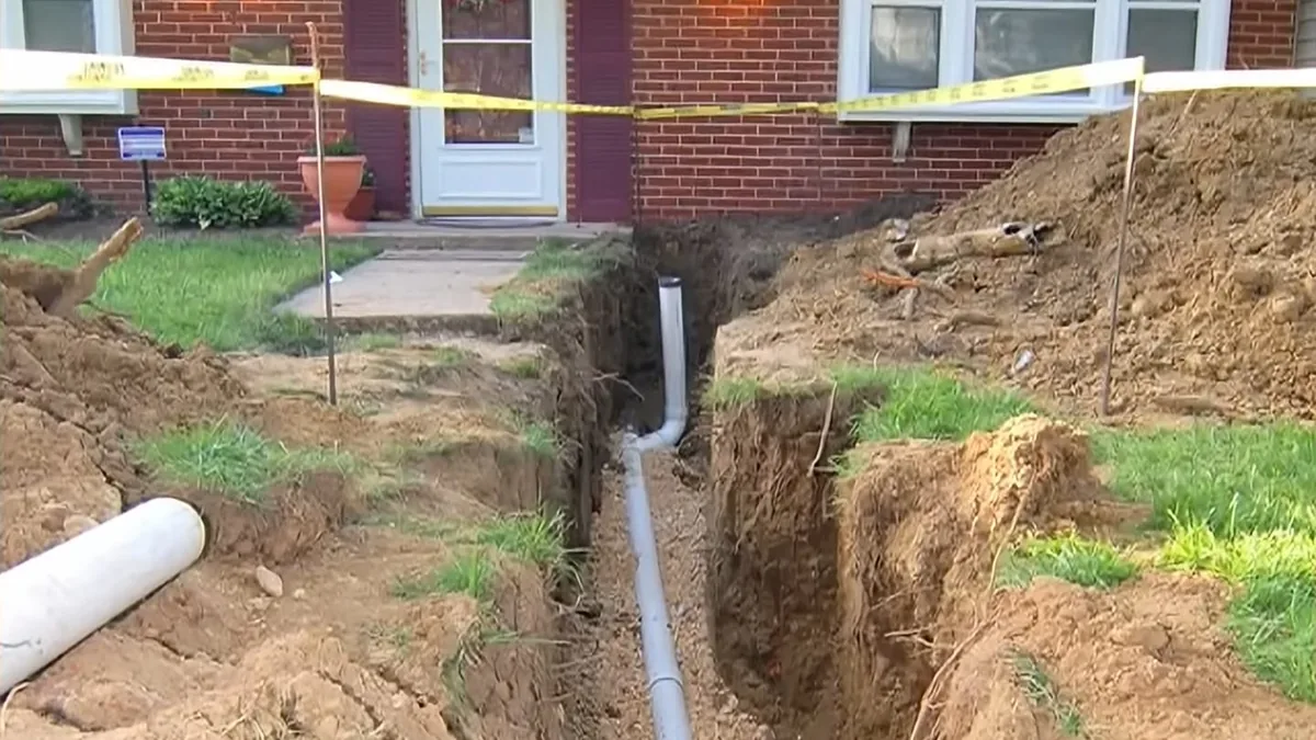 Open trench across a front yard with a new gray sewer pipe laid in it, in front of a brick house with caution tape
