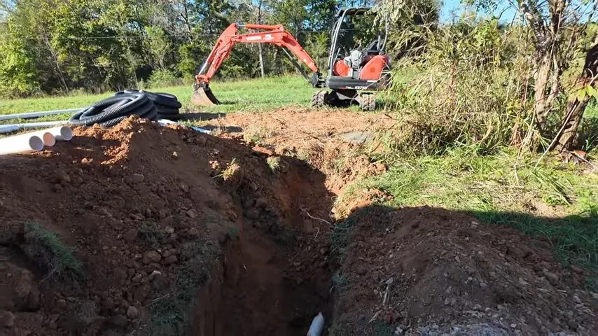 Orange mini excavator beside an open trench with lengths of black and white pipe laid in the grass