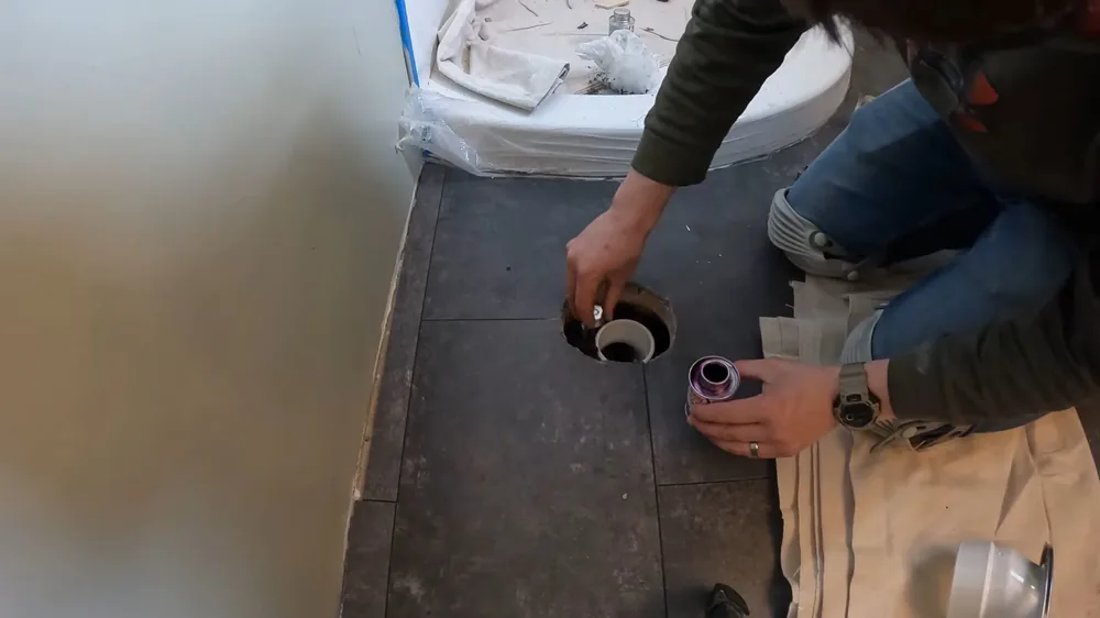Person kneeling beside a toilet flange in a floor, reaching into the drain opening with towels laid down