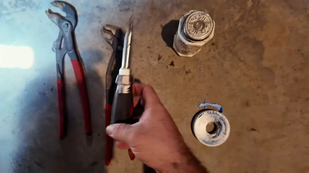 Two pairs of red-handled pliers and two threaded pipe fittings on a concrete floor, with a hand gripping one tool
