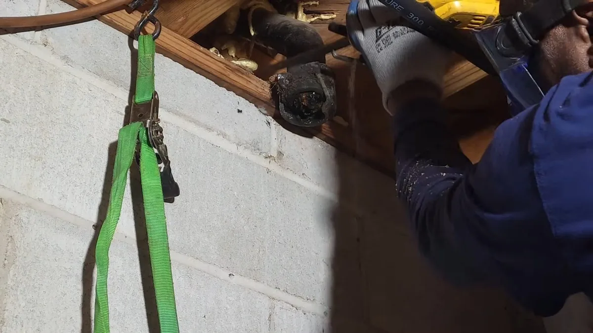 Plumber in a crawl space cutting an exposed cast iron pipe with a power saw, a green strap hanging beside him