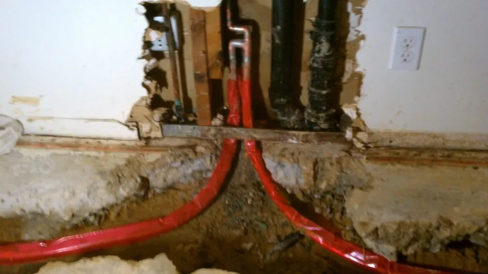 Red PEX lines curving out of a channel cut into a wall, beside dark pipes