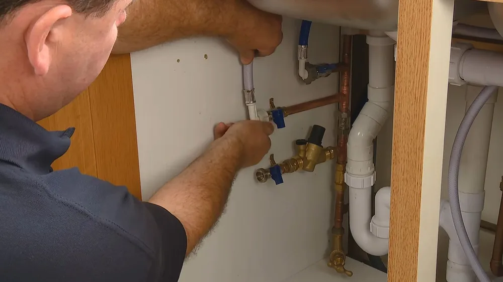Technician reaching under a kitchen sink to connect a supply line and blue-handled valve to copper pipe