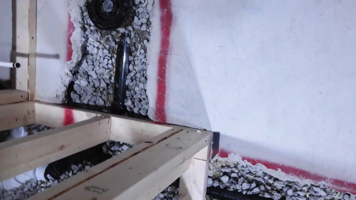 Wall cavity packed with gravel around a black drain pipe, with red tape marks on the framing