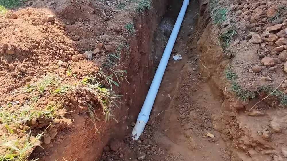 White PVC sewer pipe laid in a narrow red-dirt trench with grass along the edges