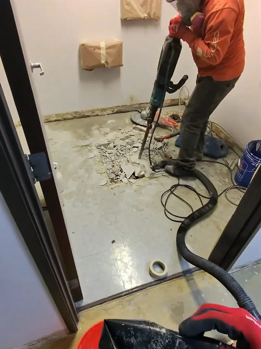 Worker in an orange jacket breaking up a tiled bathroom floor with a hammer drill, with a vacuum hose and debris on the floor