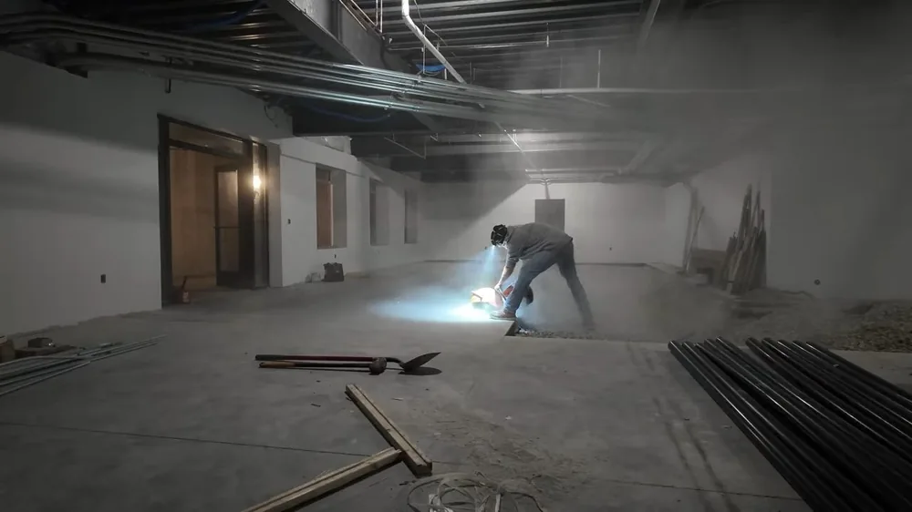 Worker cutting a line in a concrete floor with a saw, sending light and dust across an unfinished interior
